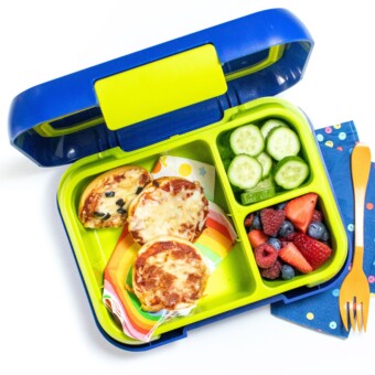 Colorful kids lunch box with bagel bites, cut cucumber and chopped fruit on a white counter with a blue napkin and orange fork.