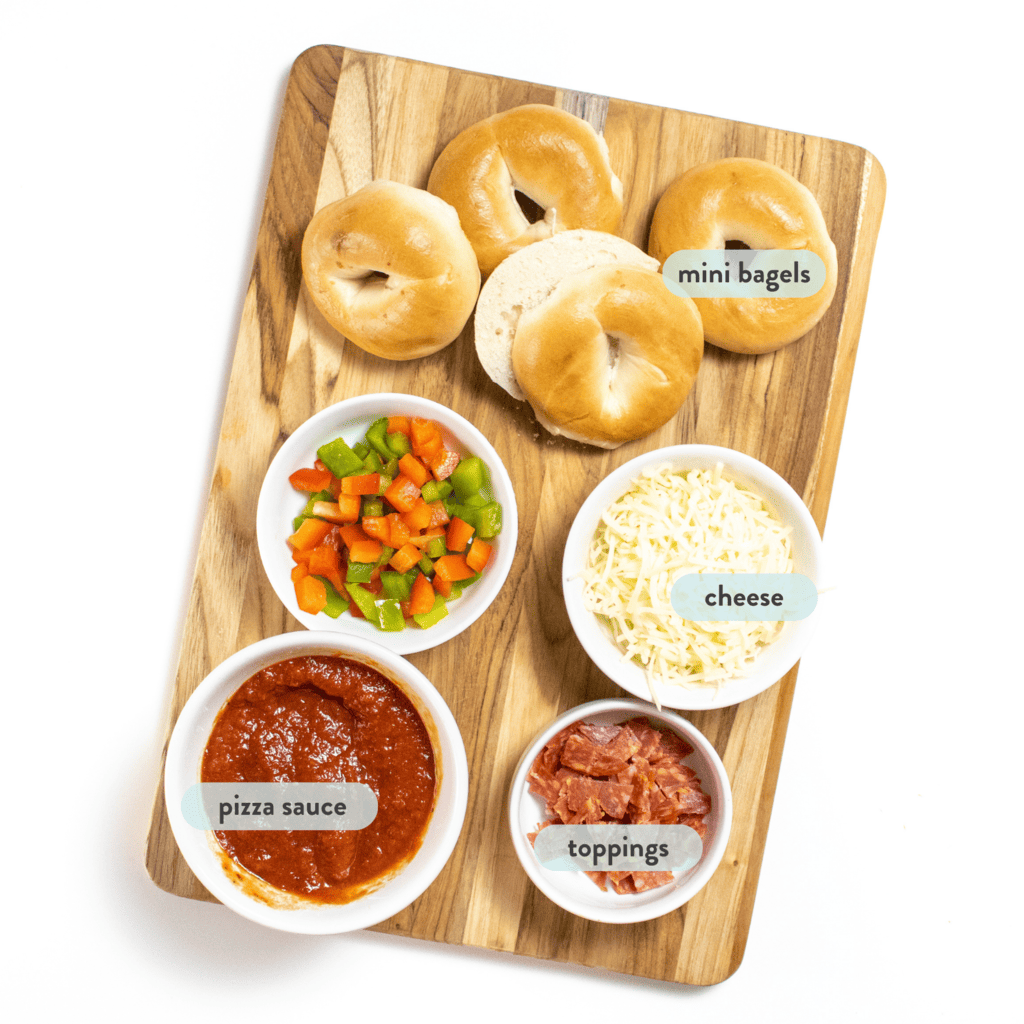 Ingredients for bagel bites on a cutting board with graphics labeling them in blue against a white background.