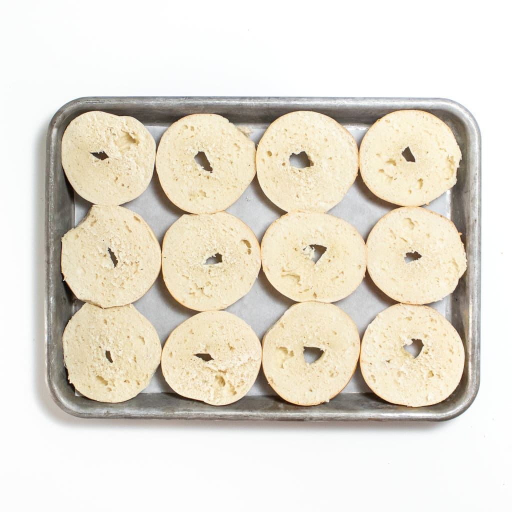 Silver baking sheet on a white counter with cut in half mini bagels on it.
