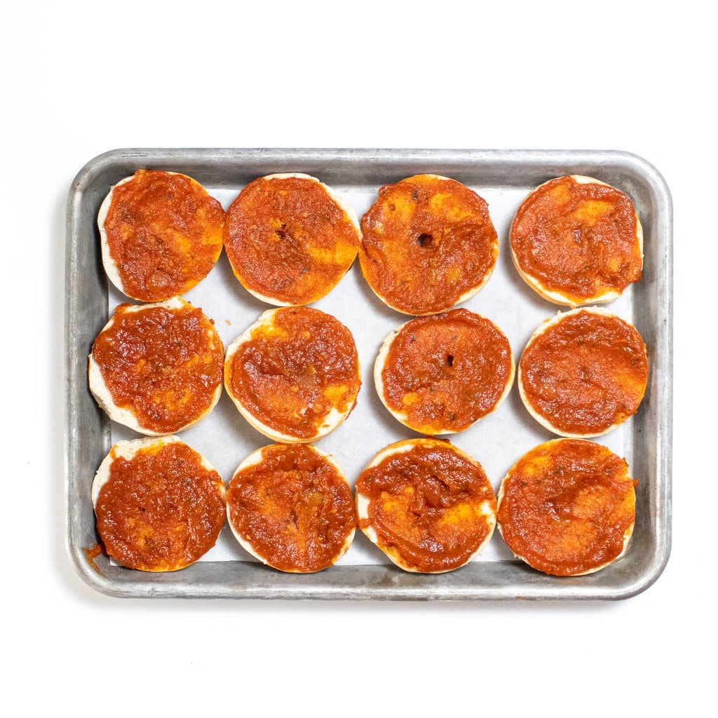 Mini bagels with pizza sauce on a baking sheet.