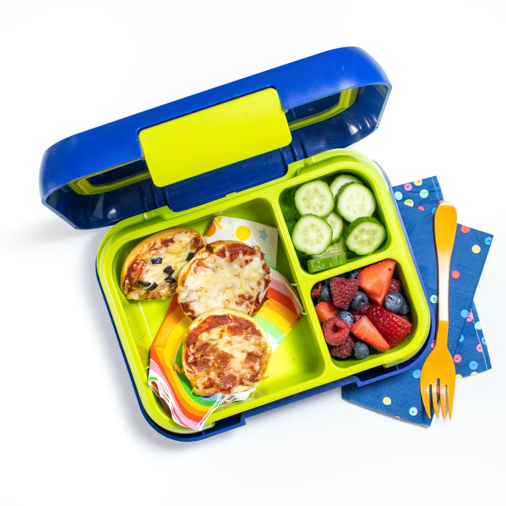 Colorful kids lunch box with bagel bites, cut cucumber and chopped fruit on a white counter with a blue napkin and orange fork. 