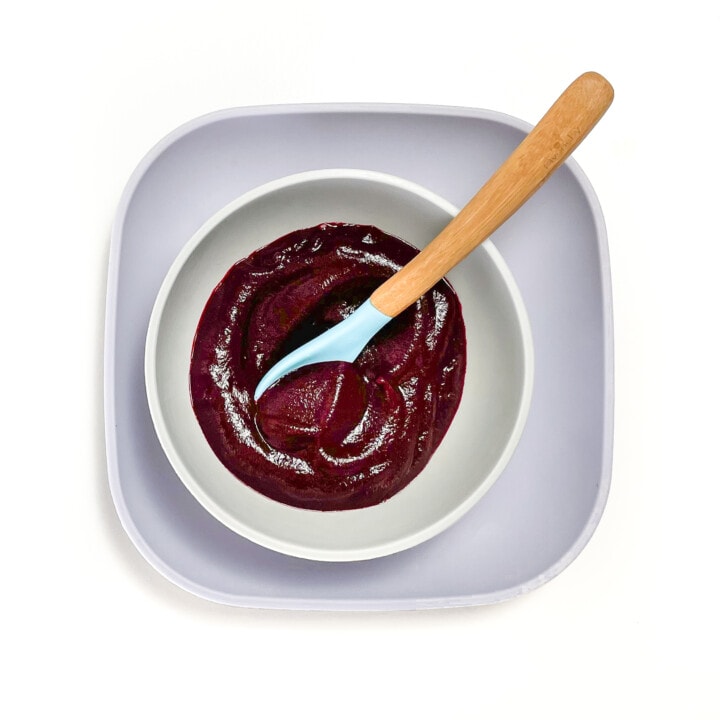 Purple and gray baby bowl filled with a bright dark red beet puree with a wood and blue spoon resting inside.