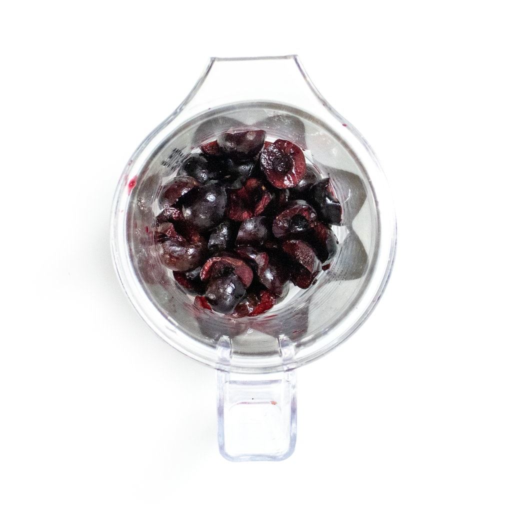 Clear blender with pitted cherries sitting on a white counter.