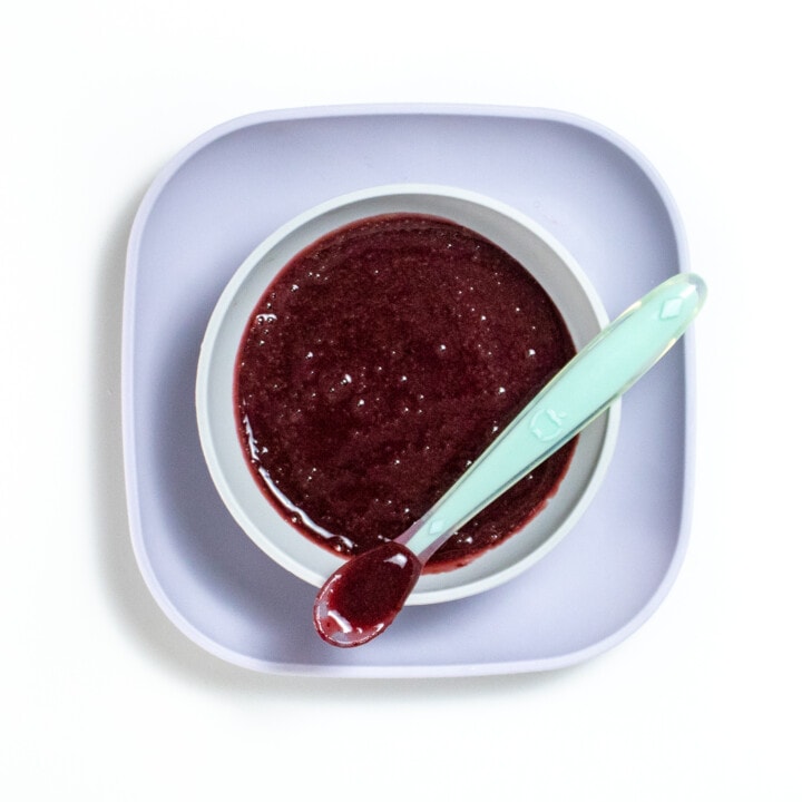 Dark red cherry puree inside of a gray baby bowl sitting on a purple baby plate.