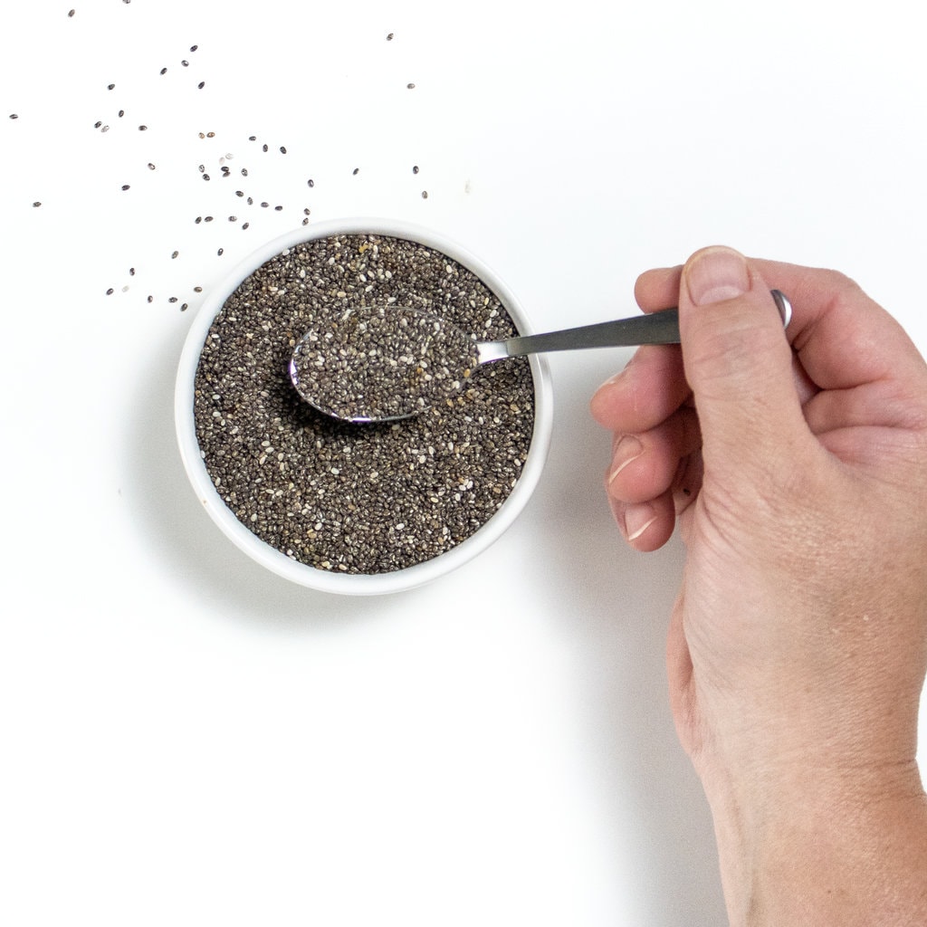 Small white bowl of chia seeds, with a hand holding a spoon full of chia seeds.