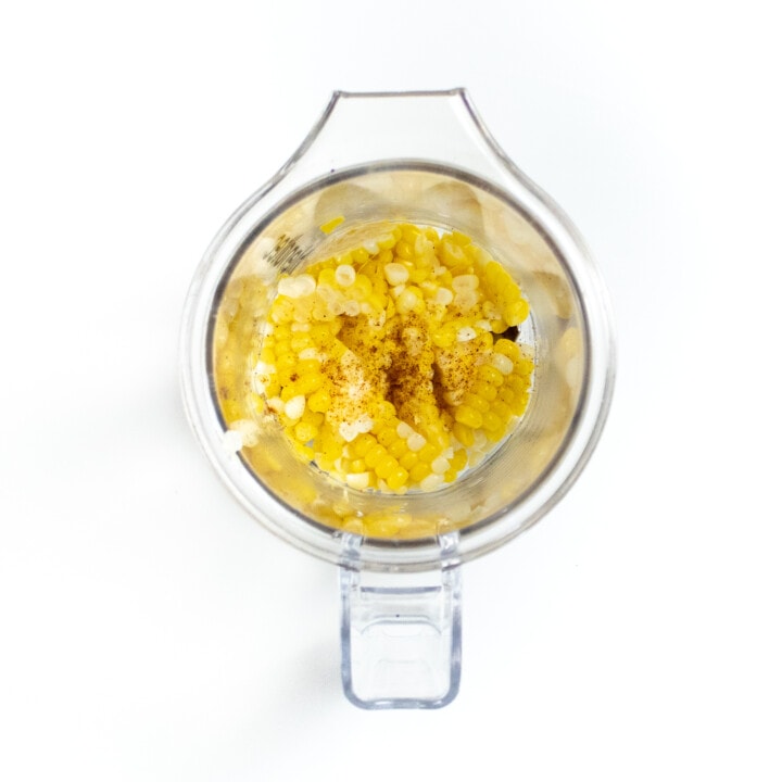 A clear blender with corn kernels and a pinch of paprika against a white background.
