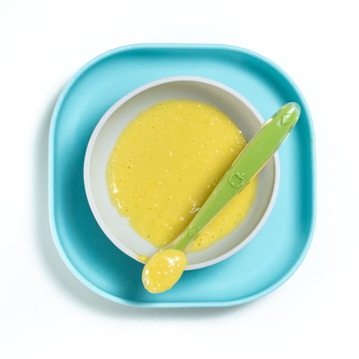 Blue baby plate with a gray baby bowl with pureed corn inside with a green spoon resting on top with puree in it.