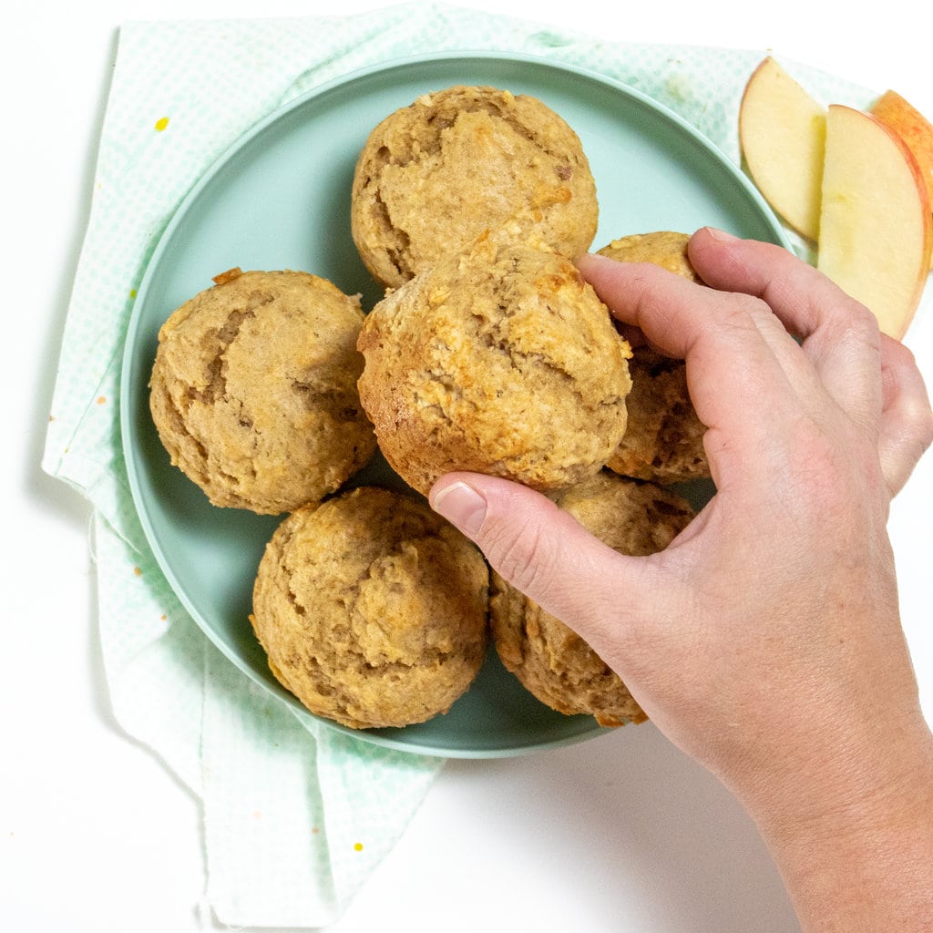 Teal kids plate with a stack of apple muffins and a hand reaching out to pick one up.