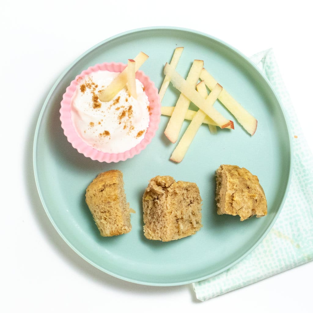 Teal blue plate with a cut up muffin, side pink bowl full of yogurt and sliced apples showing how to serve apple muffins to toddlers and kids. 