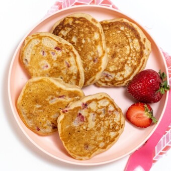Stack of strawberry pancakes on a pink kids plate with a pink fork resting on a pink napkin.