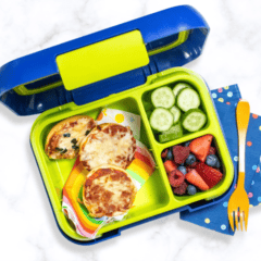 A kids blue and green lunch box filled with pizza bagel bites, cut fruit and sliced cucumbers. the bento box is sitting on a white marble counter.
