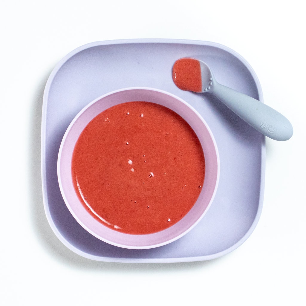 A purple baby plate with a purple bowl full of a bright pink plum puree inside, with a gray baby spoon resting on the plate.