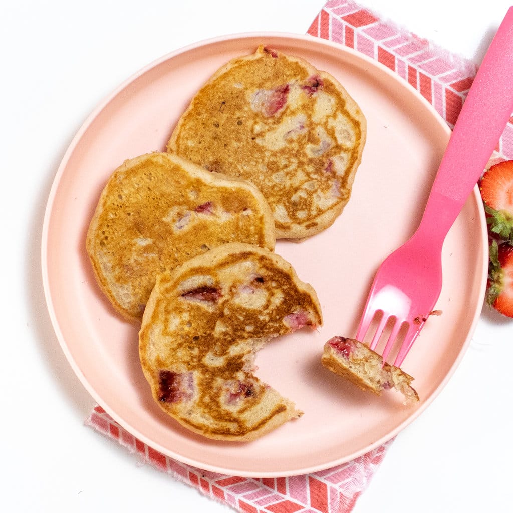 Kids pink plate with a small stack of strawberry pancakes and a pink fork cutting into one of the pancakes, all on a white counter with pink napkin.
