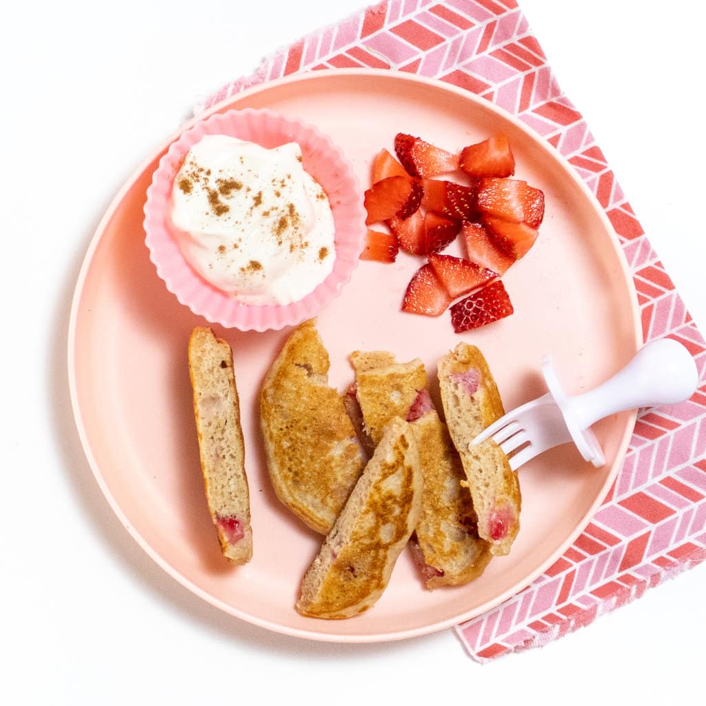 Pink kids plate full of cut up strawberry pancakes, a yogurt dip in a pink container and few chopped strawberries.