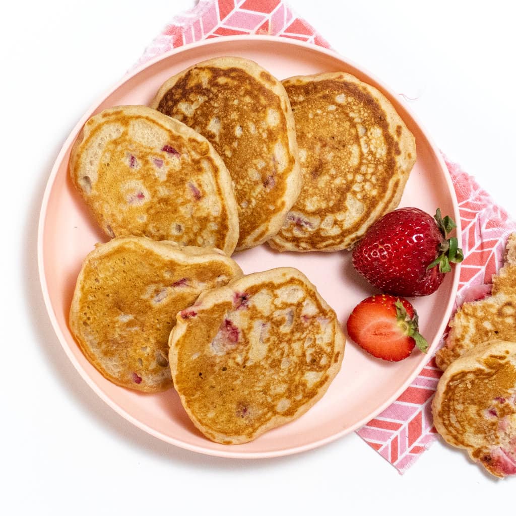 Pink kids plate with several strawberry pancakes fanned out with a few cut strawberries on the side. 