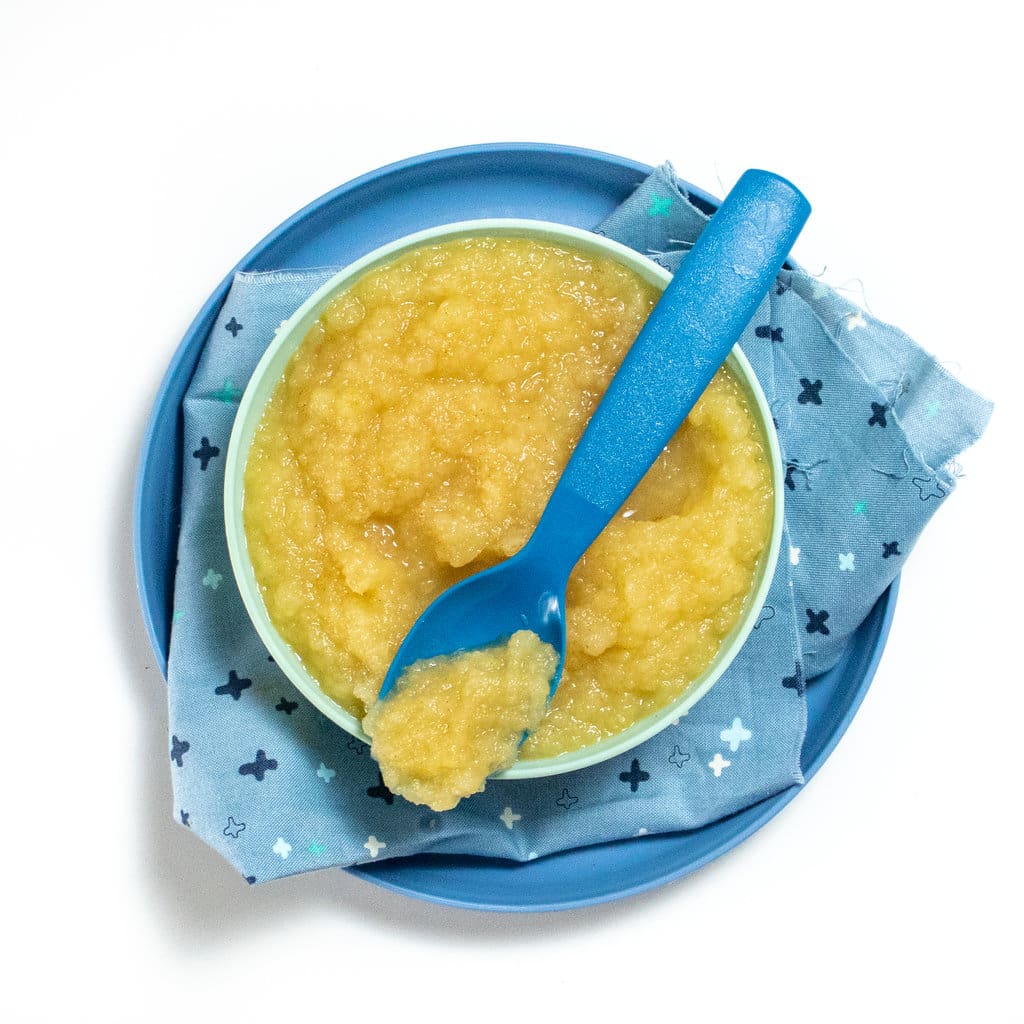 Blue kids bowl on a blue plate and blue napkin filled with homemade applesauce with spoon resting on top.