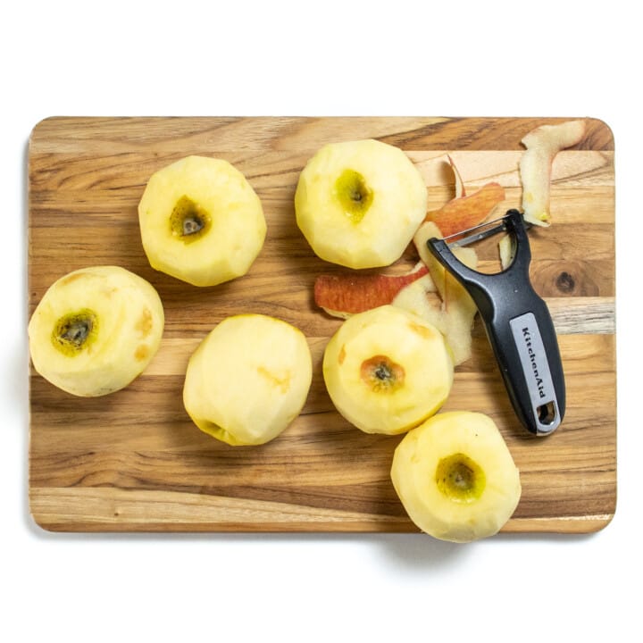Wooden cutting board with peeled apples on top with a peeler.