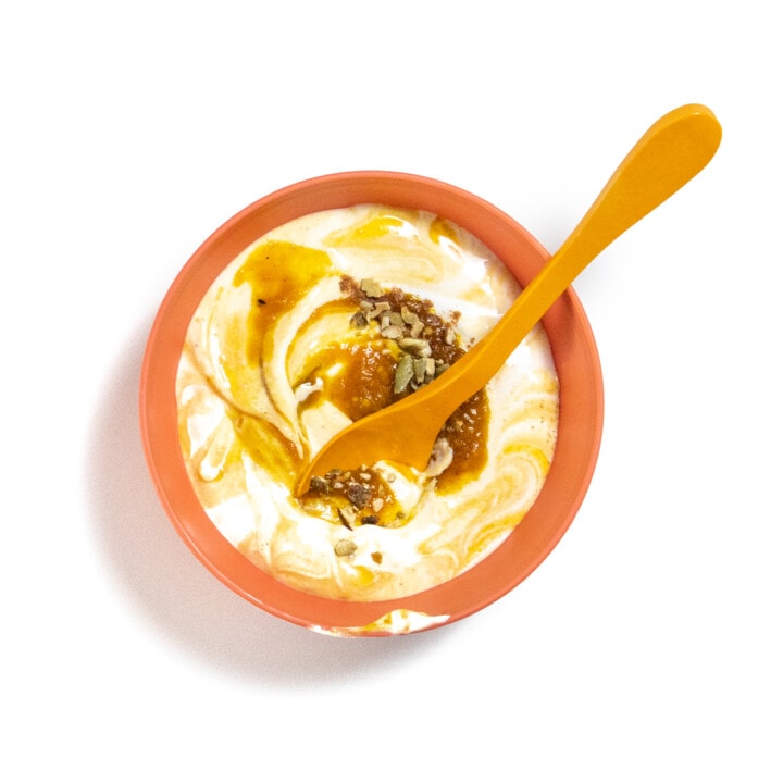 Orange kids bowl on a white counter full of yogurt with pumpkin puree and chopped nuts on top, with an orange spoon resting on top.