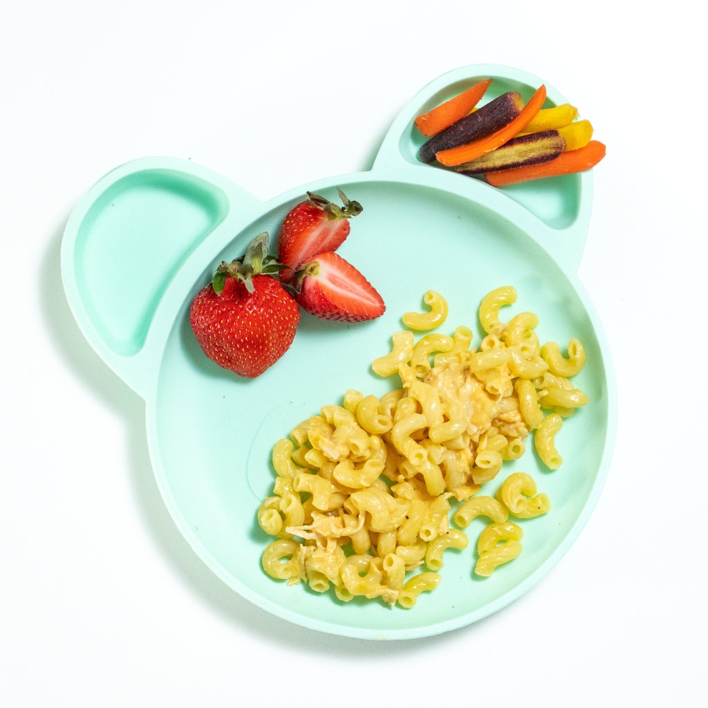 Blue toddler plate with chicken, mac & cheese, strawberries, and cut carrots on a white counter.