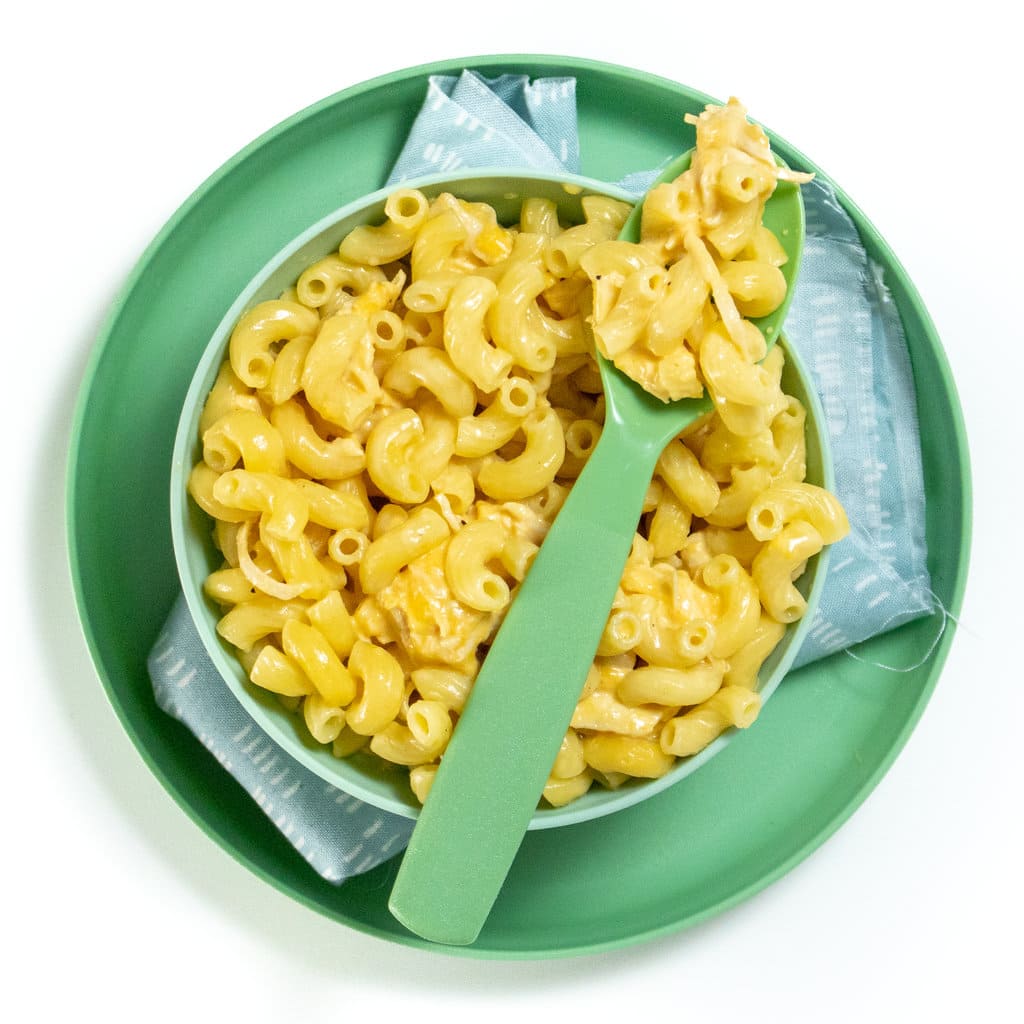 A green kids bowl and play with a blue napkin, inside the bowl is a creamy yogurt, chicken mac & cheese for kids.