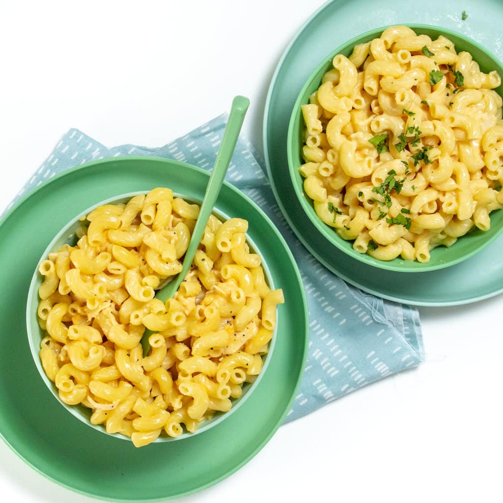 Two green kids bowls, full of macaroni and cheese with chicken with a green spoon resting inside in a blue napkin.