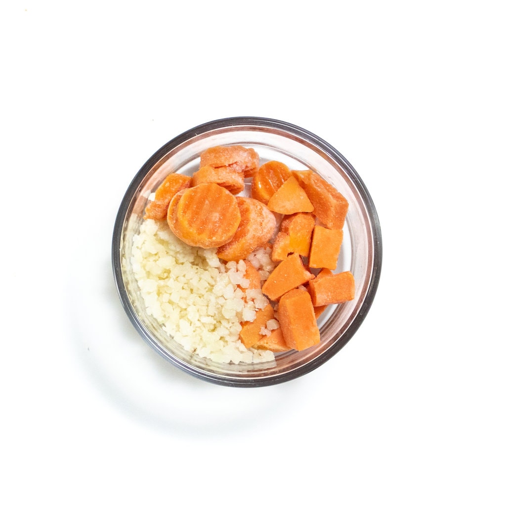 Small glass bowl, full of carrots, cauliflower and sweet, potatoes, ready to be microwaved on the white countertop.