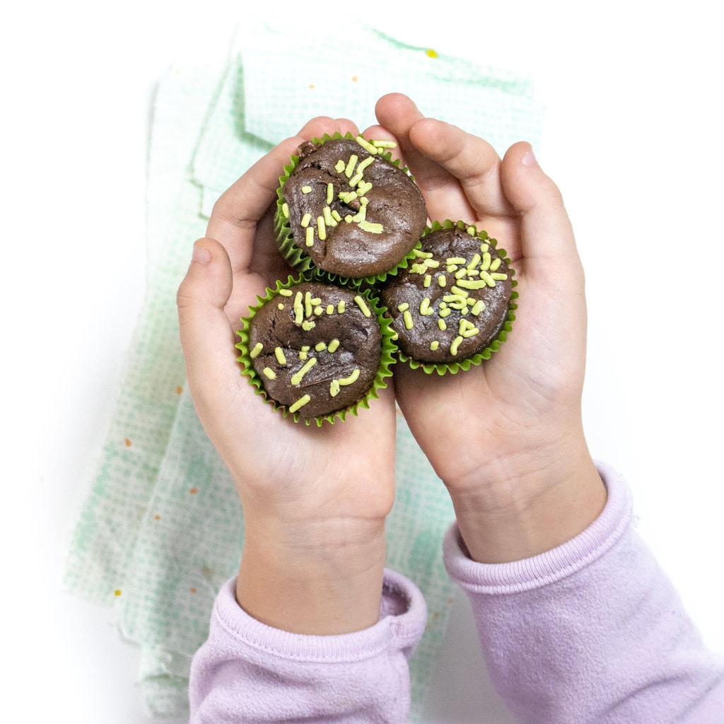 Two small kids, hands holding mini avocado, brownie bites with green sprinkles over a green and blue napkin on a white countertop.