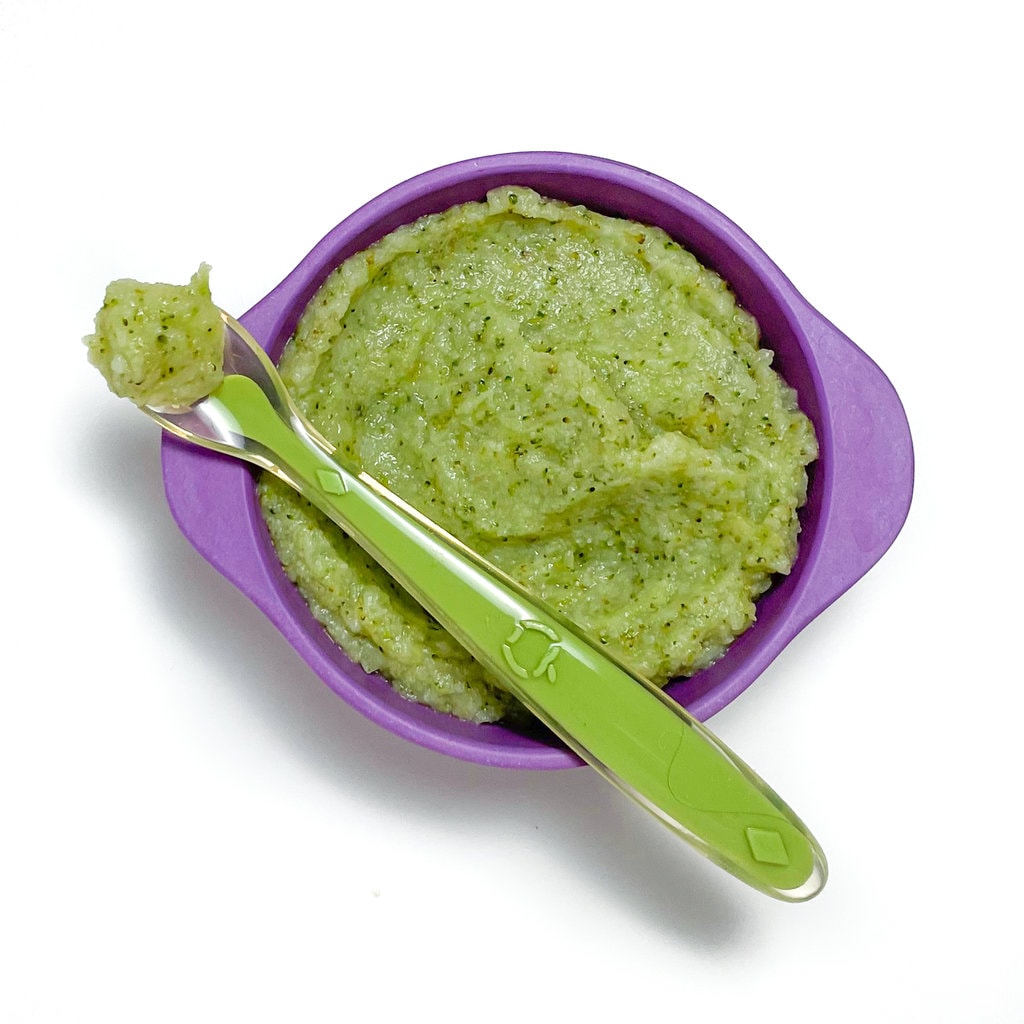 A purple baby bowl with a green baby spoon with a broccoli, cauliflower, asparagus purée.