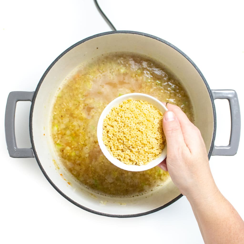 A large gray stockpot with the base for chicken and star soup with a hand, holding a small white bowl with chicken pasta over the simmering stock.