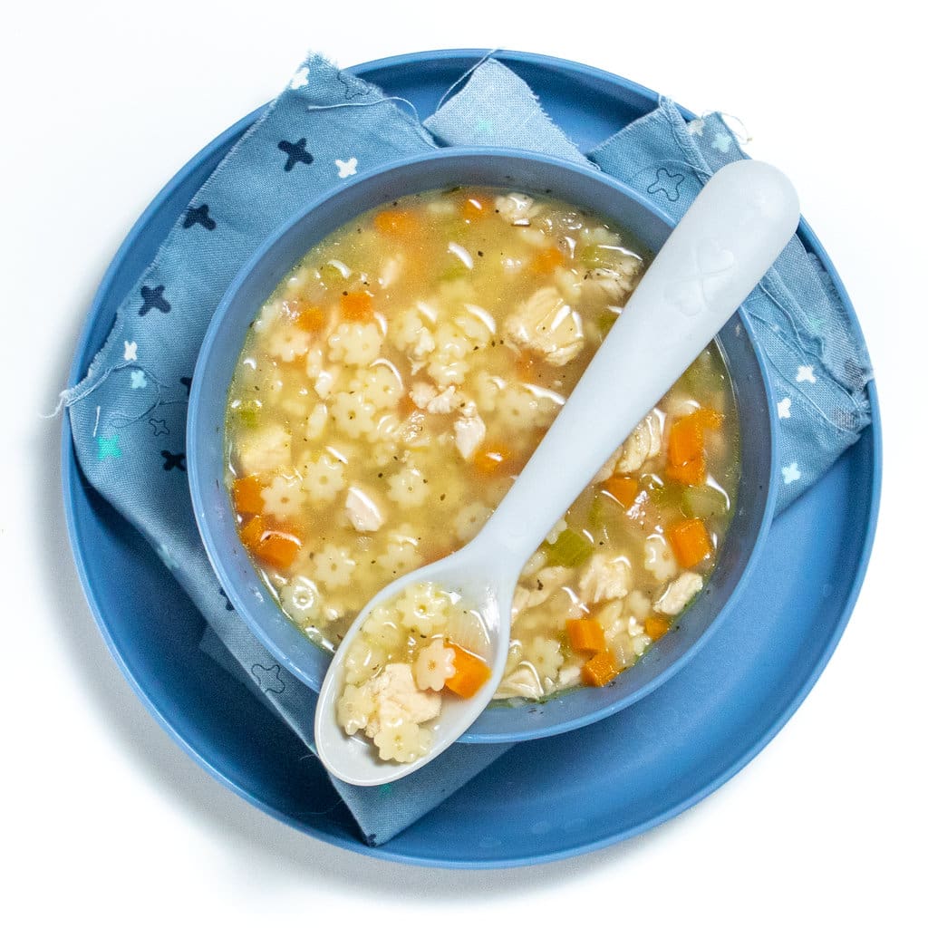 A blue plate and bowl for kids, full of chicken and star soup with a blue spoon resting on top with*pasta, shredded, diced chicken, and carrots.