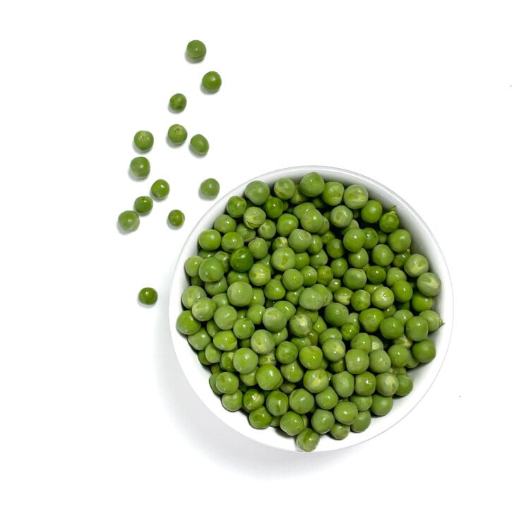 Green peas in a white bowl, spilling out on a white countertop.