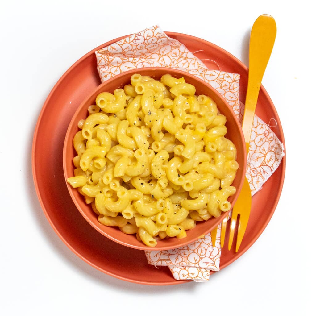 A pink plate and bowl with a colorful napkin and orange fork with three veggie mac & cheese on top.