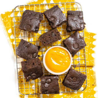 A wire rack with sweet potato brownies, cut into squares with a small bowl of sweet potato purée on top of a yellow napkin and a white countertop.