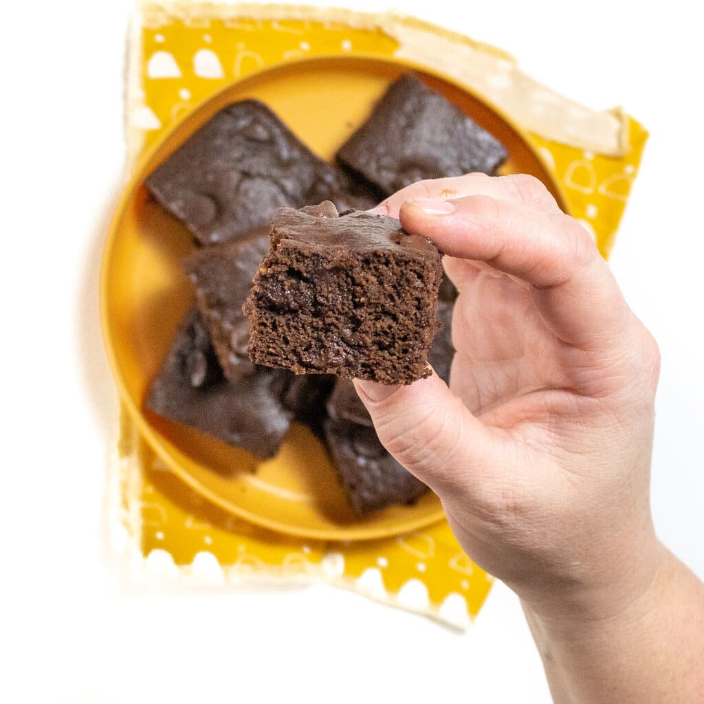 A hand holding a square cut of sweet potato brownies over a mustard, yellow plate, full of more sweet potato brownies, and a yellow napkin on a white countertop.