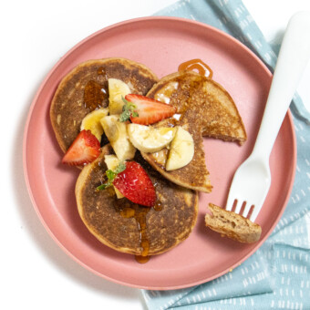 Pink kids play with three golden brown whole wheat pancakes with chopped bananas and strawberries, on top with a blue napkin and white fork.