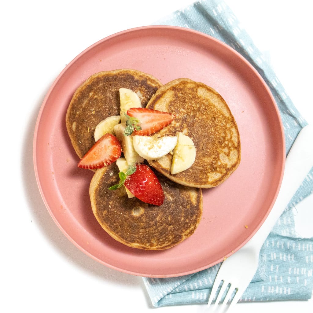 Hey kids, pink plastic plate on a white countertop with a stack of three whole wheat pancakes with banana and strawberries, with a fork and blue napkin on the countertop.