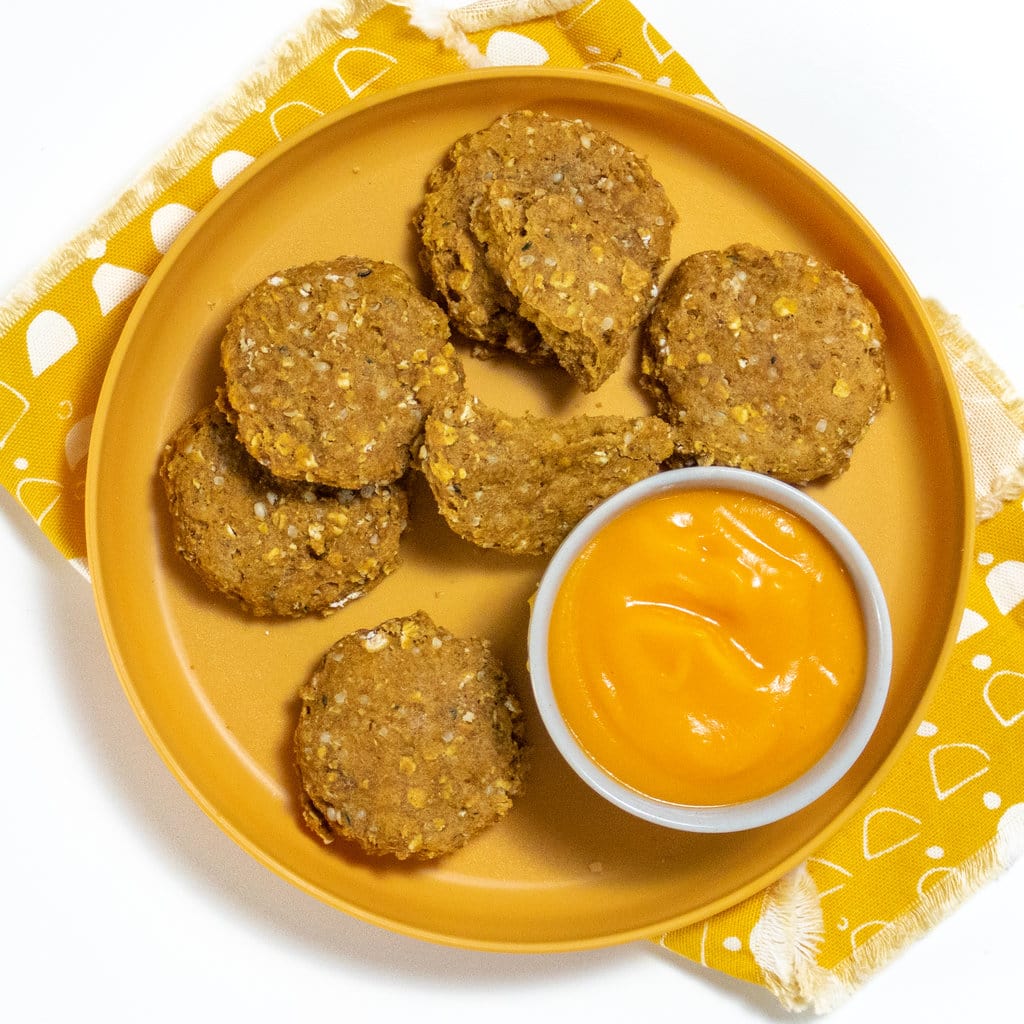 Mustard color, kids plate on a mustard color napkin on a white countertop, the plate has baby cookies stacked on top and a small bowl of sweet potato purée.