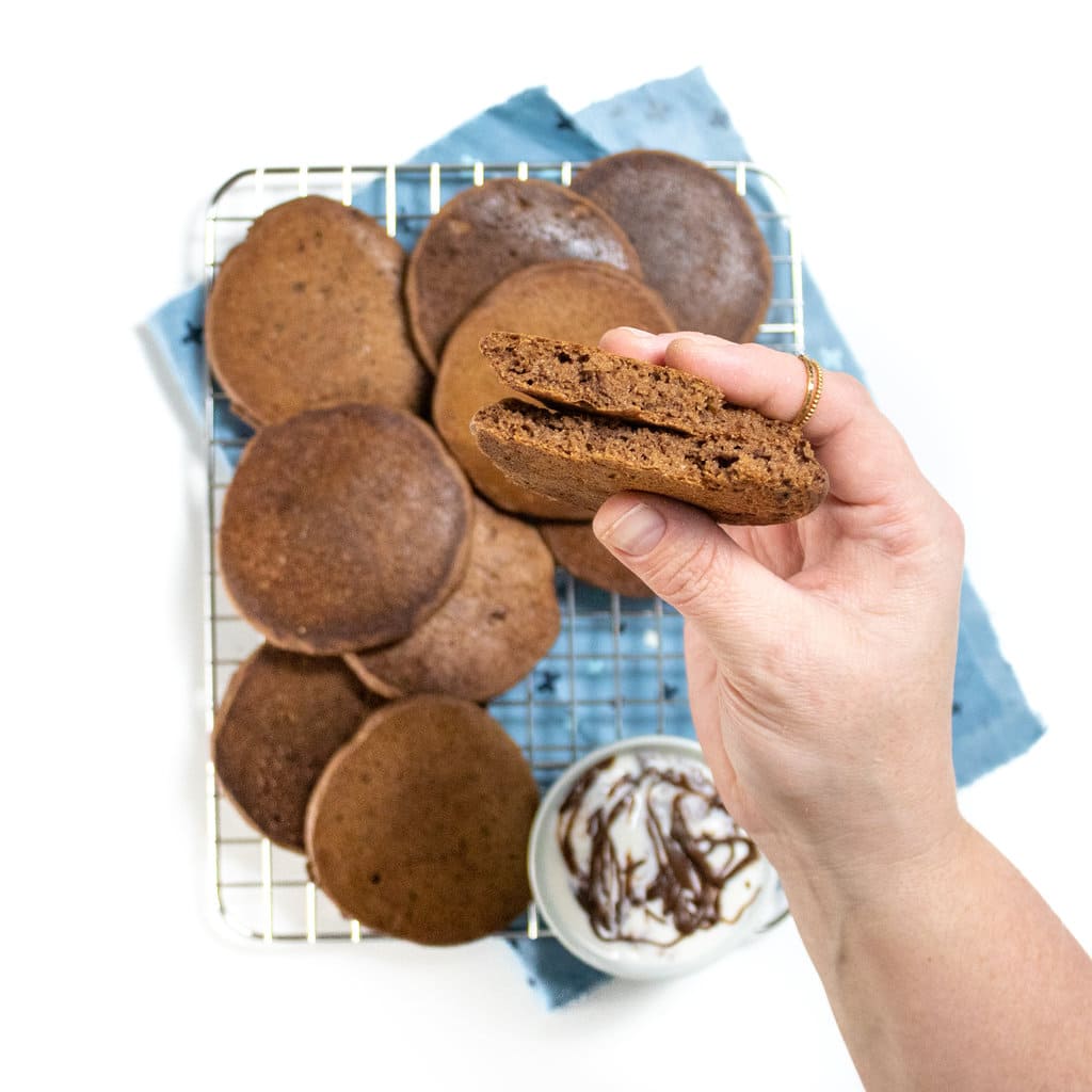 Cooling rack on a countertop with the blue napkin with a bunch of cooked chocolate pancakes in the hand, holding a chocolate pancake over top that has been ripped in half.