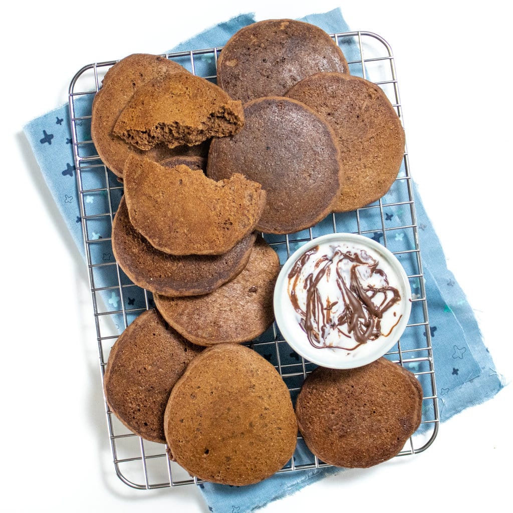 A cooling rack and a white countertop with a dozen round small cooked chocolate pancakes, and a small bowl of yogurt with chocolate sauce and a blue napkin.