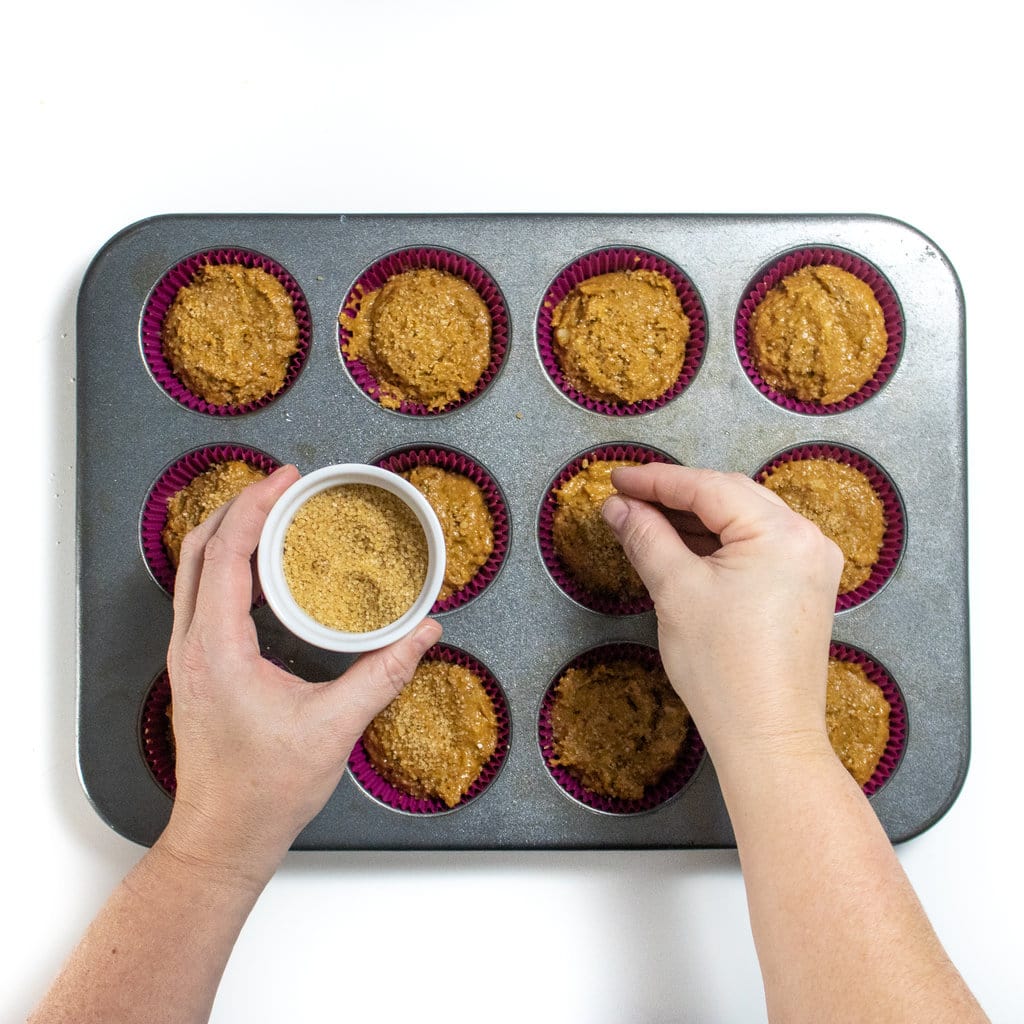 Silver muffin pan with gingerbread muffins with hands sprinkling sugar on top.
