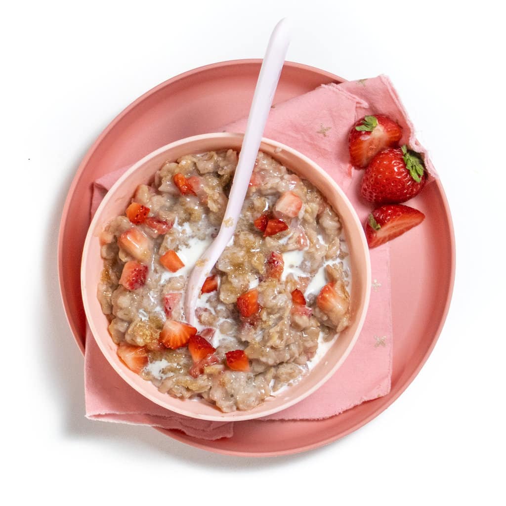Pink spoon rusting in a bowl of strawberry oatmeal with chunks of strawberries in a pink bowl and on top of a pink plate on a white marble countertop with a few strawberries scattered around.