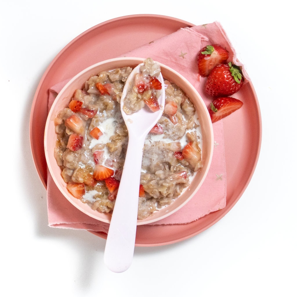 A pink spoonful of strawberry oatmeal with strawberry chunks resting on top of a pink bowl of strawberry oatmeal and a pink plate with a few strawberries on top of a white counter.