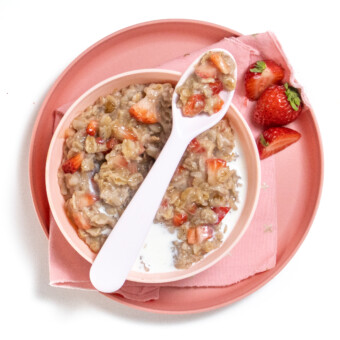 Pink kids, bowl and plate with pink napkin, the bowls, full of strawberry oatmeal chunks of strawberries, with a pink spoon, resting on top and strawberry scattered about on a white countertop.