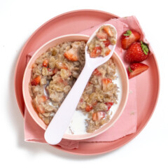 Kids bowl and plate that are pink with strawberry oatmeal inside with cream and a pink spoon resting on top with a few strawberries scattered around.