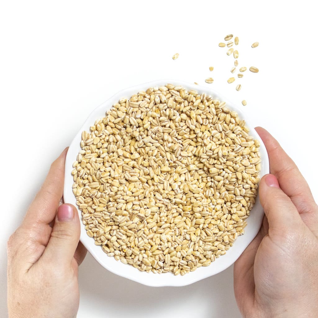 Two hands holding a white bowl, full of dry barley over a white countertop.