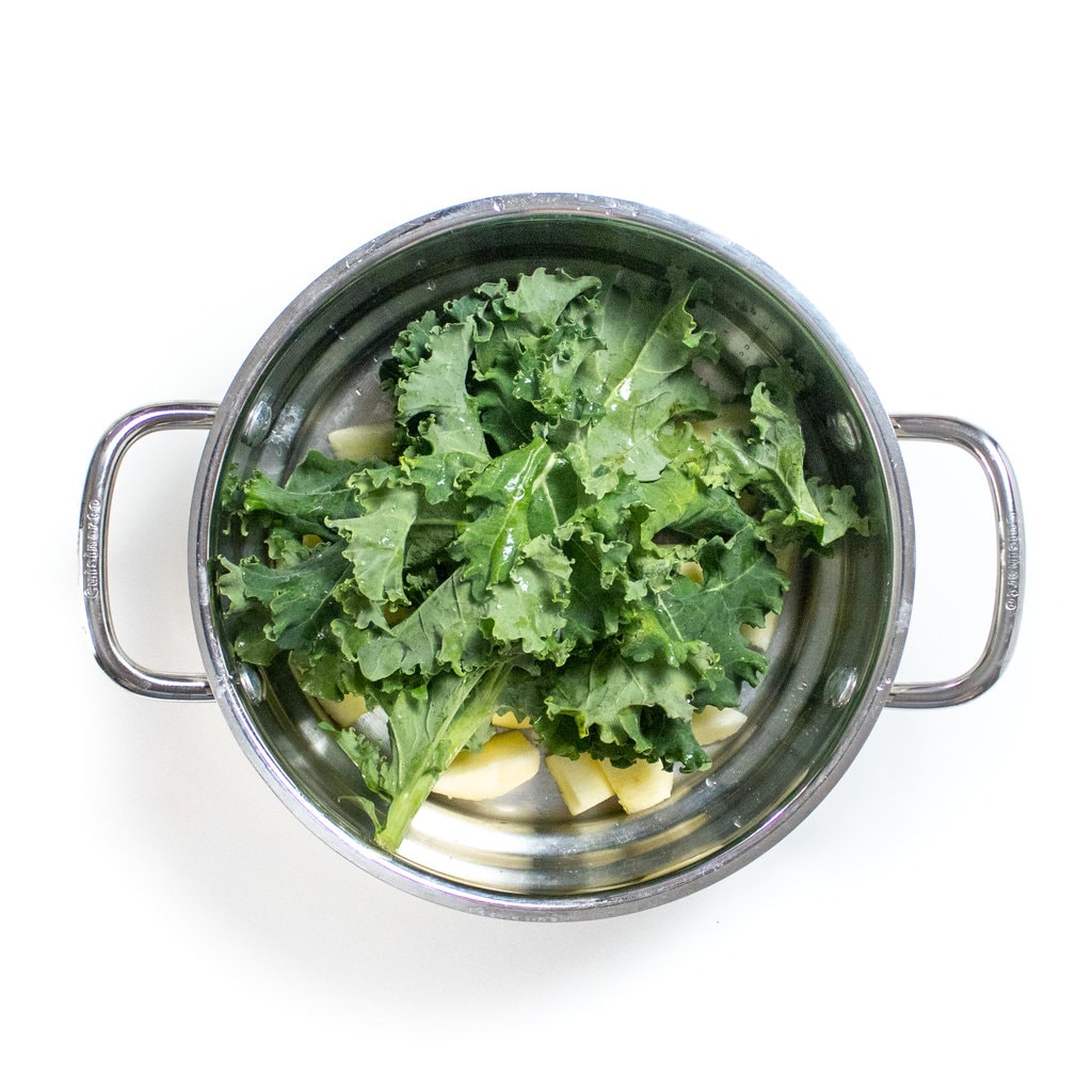 A silver steamer basket, full with kale and peeled and sliced apples.
