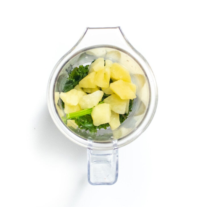 A clear blender with steamed kale and apples on a white countertop.