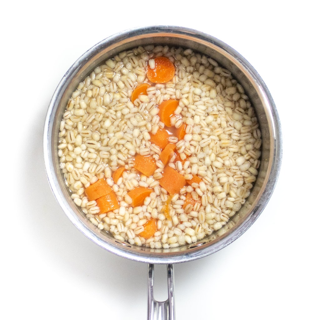Small silver saucepan over a white countertop with cooked barley and carrots.