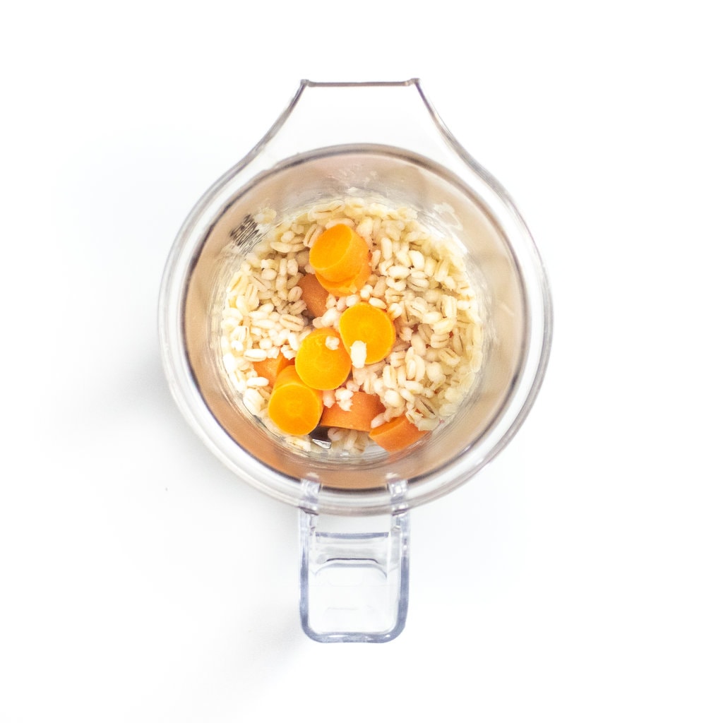 Cook, barley and carrots in a clear blender on a white countertop.