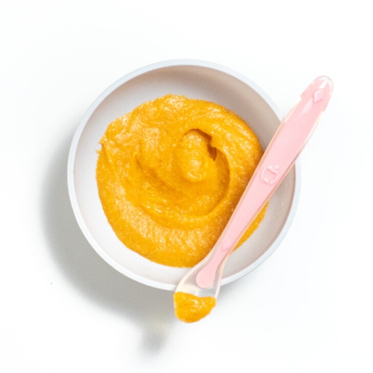 Great baby bowl on a white tank top full of carrot and barley purée with a pink spoon resting on top full of a purée.